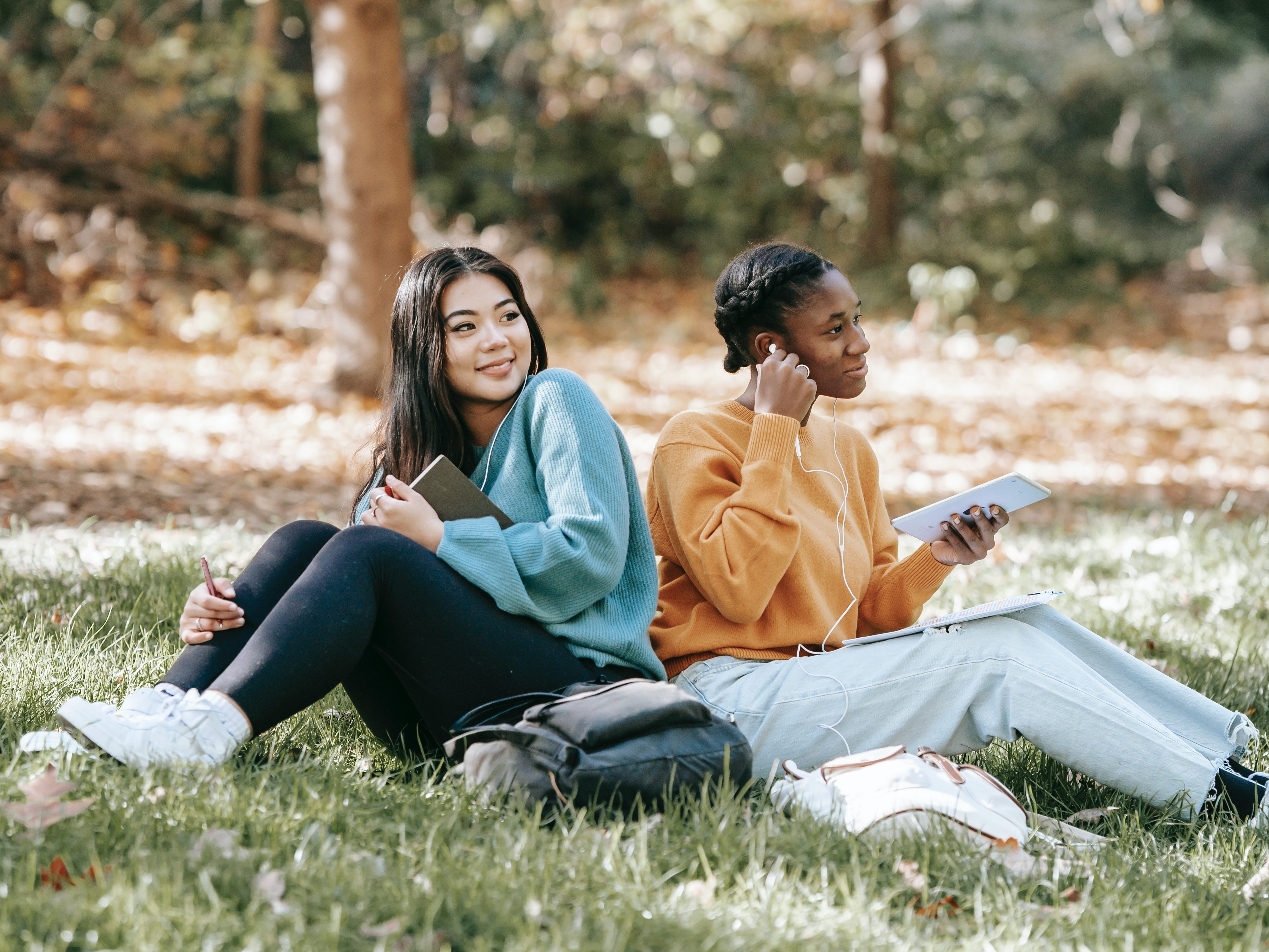 Two young women are sitting in the grass of a forest. They look to the distance with curiosity and joy.